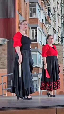 Two women in traditional flamenco attire, black skirts with red accents and red bolero jackets, stand on a wooden stage. The woman on the left, with blonde hair pulled back, holds a white fan and is in the process of opening it. The woman on the right, with dark hair, holds a red fan that is partially open. They appear to be performing a flamenco dance. The background shows a brick building with balconies and windows, suggesting an outdoor urban setting.