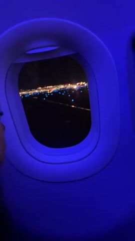 The video shows a view from an airplane window at night. The cabin interior is illuminated with blue light. Outside the window, a runway is visible with lights lining the edges and runway markers. The plane appears to be taxiing or preparing for takeoff, as indicated by the runway lights and the darkness outside. A screen is visible on the right side of the frame, displaying information. The camera is positioned to capture the view through the oval-shaped window.