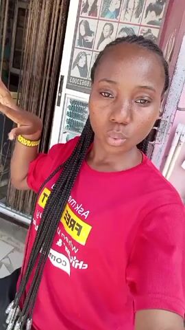 A young woman is standing in front of a beauty salon. She is wearing a red t-shirt, has long black braids, and is wearing a yellow beaded bracelet. She makes a grumpy face, then smiles and sticks her tongue out. The door to the beauty salon is visible behind her and has a poster showing different braided hairstyles. A poster of a Chelsea soccer team can be seen beside the salon door.