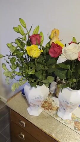 A vertical video shows two vases of flowers on a wooden dresser against a white wall. The vase on the left holds red and yellow roses, with green leaves and lily buds. The vase on the right contains white, pink, and red roses, also with green leaves and lily buds. To the right of the vases, a table lamp with a patterned shade sits on the dresser. Various small items, including hairbrushes and containers, are visible on the dresser. The camera pans slowly from left to right across the scene.