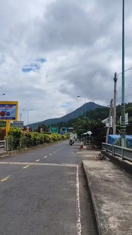 The video shows a view from a road. There is a mountain in the background, covered in trees. The sky is cloudy. There are some signs on the side of the road, and a few motorcycles driving on the road. There is also some vegetation along the side of the road.