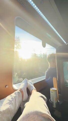 The video shows a first-person perspective from inside a moving train, looking out a window. The sun is shining brightly, casting a warm glow over a forest landscape. In the foreground, a person's legs are visible, propped up and wearing white socks and gray sweatpants. To the right of the frame, a can of what appears to be a beverage sits in a cup holder next to a headrest. In the background, another passenger with blond hair is visible, looking out the window. The train's interior features a wooden trim along the window frame and LED lighting above. The view outside the window is mostly trees and sky, with the sun creating a lens flare effect. The video is shot in landscape orientation and appears to be a short clip, likely a few seconds long, capturing a serene moment during a train journey.