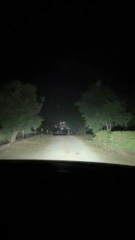 The video shows a first-person perspective of driving down a dirt road at night. Headlights illuminate the path ahead, flanked by trees and sparse vegetation. The road appears to lead towards a dimly lit structure in the distance. There are lights visible along the side of the road, possibly streetlights or pathway lights. The overall atmosphere is dark and somewhat rural.