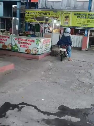 A person wearing a blue jacket, white helmet, and light blue pants rides a motorcycle. The person is sitting on the motorcycle, facing away from the camera. The motorcycle is parked in front of a food stall. The stall has a sign that says "ES KELAPA MUDA ES CAMPUR PENTOL" and "BU GİYARTİ JOK PASAR PLE". There is another person standing behind the counter of the stall, preparing food. The environment appears to be an outdoor market or a street with several stalls. The ground is paved with concrete and has some wet patches. The overall lighting suggests it is daytime, possibly overcast.