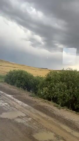 This media file captures a view from inside a vehicle, looking out at a rural landscape. The scene is dominated by a vast, dry, golden-colored field that stretches towards a distant horizon marked by rolling hills. The sky above is dramatic, filled with dark, heavy storm clouds, suggesting an approaching or ongoing weather event. A dirt road with visible puddles and gravel runs along the foreground, indicating recent rain. Lush green bushes and trees line the edge of the road, contrasting with the arid field.

The activity taking place is implied to be a journey or travel by vehicle through this natural setting. The time of day appears to be late afternoon or early evening, given the lighting and the mood created by the storm clouds. The weather is clearly inclement, with a palpable sense of impending rain or a recent downpour. The location context points to Karagayl, Kazakhstan, which aligns with the description of a vast, open, and somewhat arid landscape. There are no visible people or specific objects of note in the frame, only the natural elements and the road. The overall emotion conveyed is one of vastness and the power of nature, perhaps with a touch of somberness due to the stormy sky. No text is visible in the media file.