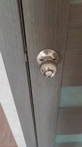 This media file shows a close-up, slightly angled shot of a person's hand opening a grey wooden door. The door has a metallic doorknob. The hand, appearing to be an adult's, grips the doorknob and turns it to the right. The background shows a textured wall and the edge of the door frame. The lighting suggests an indoor setting, possibly during the daytime. No visible text is present in the media file. The activity depicted is the simple act of opening a door. The focus is on the interaction between the hand and the doorknob, highlighting the motion of opening. The setting appears to be a residential or office space due to the standard door and doorknob design.