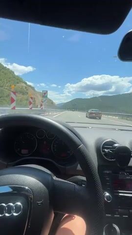 The video captures the perspective of a driver inside an Audi car traveling on a highway. The dashboard is visible, showing the steering wheel, speedometer, and radio. Outside the car, a scenic view of mountains and a blue sky with white clouds unfolds. The car is in motion, indicated by the blurred scenery and the movement of the vehicle ahead on the highway. The time stamp shows the video starts at 00:00 and ends at 00:04.