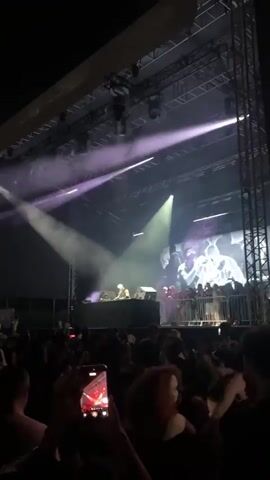 A live music performance is taking place on a stage. A DJ is behind a table with equipment, and a large screen displays visuals behind them. There is a crowd of people in front of the stage, some of whom are holding up their phones. Lighting effects, including spotlights and smoke, are being used. The scene is dark, illuminated by stage lights.