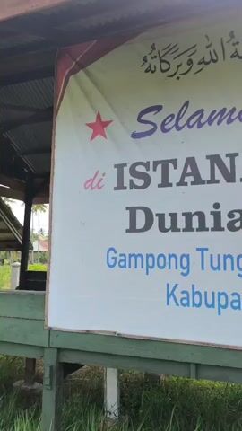 The video shows a close-up of a banner with text in Arabic and Indonesian. The Arabic text at the top reads "Assalamu alaikum wa rahmatullahi wa barakatuh," which translates to "Peace, mercy, and blessings of Allah be upon you." Below this, in purple cursive text, is "Selamat Datang," meaning "Welcome." Further down, in large, bold, black text, it says "ISTANA TRANSIT DUNIA - AKHIRAT." Below this, in blue text, is the location: "Gampong Tungkop Kec. Darussalam Kabupaten Aceh Besar." The banner is mounted on a wooden frame and is partially obscured by foliage and some rolled-up objects at the bottom. The environment appears to be outdoors, possibly in a rural area. There are no people visible in the video.