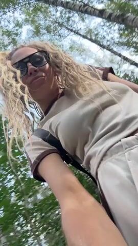 A blonde woman with long, wavy hair and large, dark-rimmed glasses is taking a selfie while walking or riding outdoors. She is wearing a light-colored t-shirt with dark stripes on the sleeves and a dark backpack. The background shows a path, trees, and greenery, suggesting a park or wooded area. The camera movement indicates she is in motion.