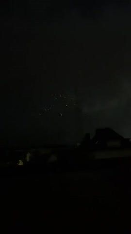 The video shows a nighttime scene with fireworks exploding in the sky. The fireworks are various colors, including orange, yellow, and white. There are also some faint lights visible in the distance, possibly from houses or streetlights. The overall mood is festive and celebratory.