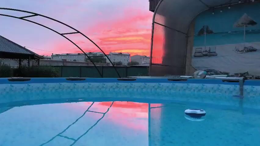 The video shows an outdoor swimming pool during a vibrant pink and orange sunset. The sky is filled with colorful clouds reflecting on the surface of the pool. In the background, there are residential buildings and a green fence. A pool cover structure is visible to the right of the frame, and a small white and blue pool cleaner or float is in the water. The scene captures a tranquil evening atmosphere.