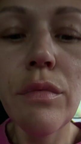 The video shows a close-up, front-facing shot of a woman's face. Her eyes are initially looking upwards and then widen dramatically as she stares directly at the camera. The focus is on her facial expression, particularly her wide eyes and slightly parted lips. The lighting is dim, with some highlights on her skin. The background is out of focus, but appears to be an indoor setting, possibly a car.