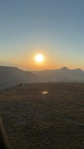 The video captures a serene sunrise over the mountainous landscape of Yeşilhisar, Turkey. It begins with a wide shot of the sun rising, casting a warm, orange glow across the horizon and silhouetting distant mountains. The foreground features a vast, dry, grassy plain.

The perspective shifts to a person viewing the scene from behind, with a hot air balloon visible in the far distance, hinting at the main activity. The video then widens again, revealing multiple hot air balloons ascending into the clear blue sky. The landscape below remains a mix of dry grasslands and possibly some cultivated fields. One prominent white hot air balloon with red and black lettering, partially visible as "TAL...", stands out against the sky, while other yellow and various colored balloons dot the horizon. The distinct sound of a hot air balloon's burner can be heard, indicating they are either taking off or actively flying. The overall scene conveys a peaceful and majestic early morning experience, characteristic of hot air balloon flights at dawn.