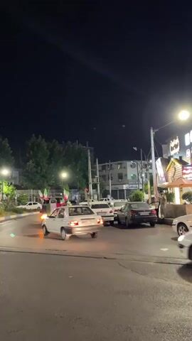 A night scene shows several cars driving on a street. Many of the cars have flags attached to them, including Iranian flags and a red flag. People are visible inside and around the cars. The street is paved, and there are buildings and trees visible in the background. Streetlights illuminate the scene. One car in the foreground appears to be moving towards the viewer. The overall impression is of a celebratory or festive event.