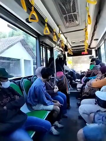 The video shows the interior of a bus with passengers seated. The camera is positioned to view the aisle and the seats on both sides. Several individuals are visible, some looking out the windows, others talking or resting. The seats are a combination of green and blue upholstery. Yellow grab handles are attached to the ceiling. A digital display screen above the windshield shows red text, possibly indicating the destination or status of the bus. Outside the windows, glimpses of trees and a structure can be seen, suggesting the bus is in motion, likely on a road with some vegetation. A fire extinguisher is mounted on a pole near the back of the bus. The overall atmosphere appears to be that of a typical public transportation journey.