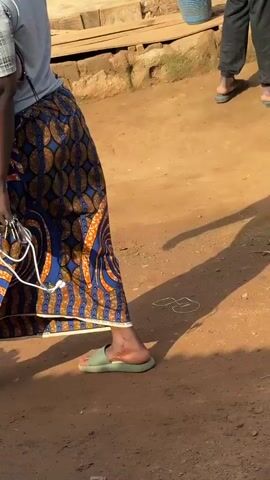 The video shows a bustling marketplace scene. The camera pans across various stalls displaying food items and produce. In the foreground, a person wearing a colorful skirt is seen holding a white wire and keys. To the left, a wooden stand holds a plate of what appears to be cooked meat. Further back, large buckets filled with grains and other food items are visible. The ground is dusty and unpaved, indicating an outdoor market. The stalls are made of wood and corrugated metal, with various goods neatly arranged. The overall impression is one of a lively and busy local market.