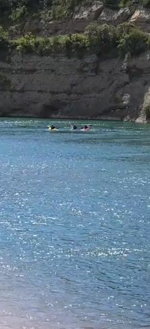 The video shows three people kayaking in a river on a sunny day. The water is a light blue color and is reflecting the sunlight. The kayaks are yellow, blue, and red, and the kayakers are each paddling. The river is flanked by steep, rocky cliffs covered in vegetation.  A power line is visible in the background. The camera is stationary.