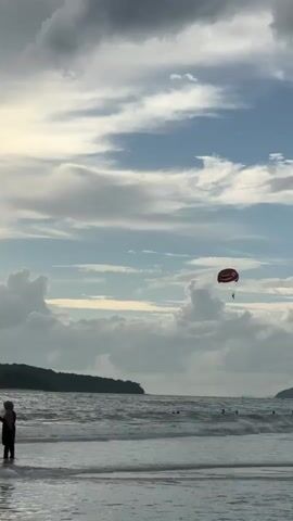 The video begins with a view of the ocean and beach, with a parasailer in the sky. As the video progresses, the camera pans to the right, showing more of the coastline and the ocean. There are several people in the water, some swimming and others standing near the shore. The sky is cloudy, but there are patches of blue visible. A small boat is seen in the distance. The video ends with a person squatting in the shallow water near the beach, appearing to be playing or looking for something.