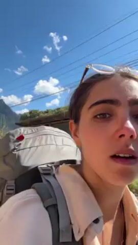 A woman with her sunglasses on her head, a large backpack, and a light pink shirt is walking on a dirt road in a rural area. She appears to be filming a selfie-style video. In the background, there are simple houses, trees, and a mountain range under a clear blue sky with some clouds. Power lines are visible overhead. The road appears to be lightly traveled and the overall setting evokes a sense of rustic travel.