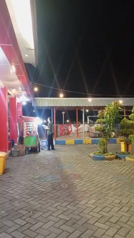 This is a vertical video taken at night. The scene appears to be an outdoor area with a paved ground made of bricks. In the foreground, there are swings with brightly colored posts. Two young boys are seated on the swings. To the right of the swings, a person wearing a green top is sitting on a swing, facing away from the camera, with another person standing behind them. In the middle ground, there are trees and other plants. A striped tent-like structure is visible behind the swings. String lights are hung across the area, casting a warm glow. To the left of the scene, a food cart is set up, and a person is standing near it. The background is dark, indicating it is nighttime. The overall impression is a lively outdoor space, possibly an amusement park or recreational area.