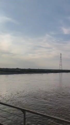 The video pans across a wide, muddy river under a blue sky with scattered clouds. On the right side of the frame, a light-colored metal railing is visible, suggesting the viewpoint is from a bridge, boat, or elevated platform. The landscape on the far bank appears to be mostly flat with trees and some structures visible in the distance, particularly a tall tower with lights. The water is choppy, indicating some movement or current. The camera movement is smooth, suggesting a steady platform. There are no people visible in the video. The lighting indicates it is daytime.