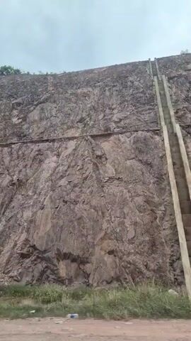 The video shows a large, steep, rocky cliff face. There are visible horizontal lines embedded in the rock, suggesting some form of man-made intervention, possibly for stabilization or excavation. Vegetation, including grass and small trees, is present at the base of the cliff and along the very top edge. The sky above is overcast. The camera appears to be positioned low to the ground, looking upwards at the cliff. There are no people or animals visible in the frame. The overall impression is of a quarry or a cut into a mountainside.