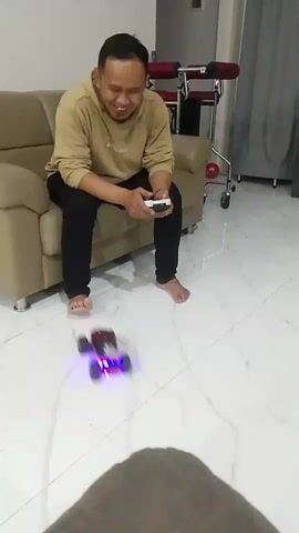 A man is sitting on a couch holding a remote control. A remote-controlled car with blue lights is on the floor in front of him. The man appears to be laughing and interacting with the car. The environment appears to be a living room with a tiled floor and a couch.