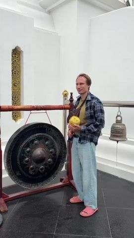A person wearing a plaid shirt, a tan shirt, light blue jeans, and pink slippers stands in front of a large, dark gong hanging on a wooden stand. The person is holding a yellow mallet in their right hand and is about to strike the gong. Another bell is visible hanging to the right of the gong. The background consists of white walls and a golden decorative element on the left wall. The floor is dark gray.