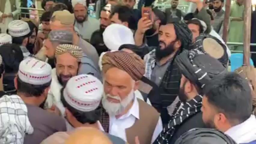 Here's a concise description of the video:

The video shows a large crowd of men gathered together. Many of the men are wearing traditional clothing, including white caps with red embroidery, turbans, and vests. Some individuals are taking photos or videos with their phones. There appears to be movement within the crowd, indicating people are pushing or trying to move through the group. The overall environment appears to be outdoors and possibly under a tent or structure based on the lighting and visible framework in the background.