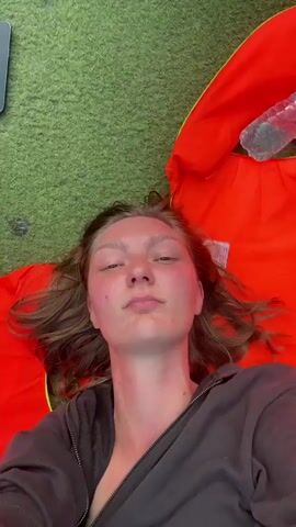 A woman lies on her back on an orange life jacket, on top of green artificial turf. A half-empty water bottle is on the life jacket. The camera pans up to show that she is on a boat moving on a green lake. Lush mountains surround the lake.