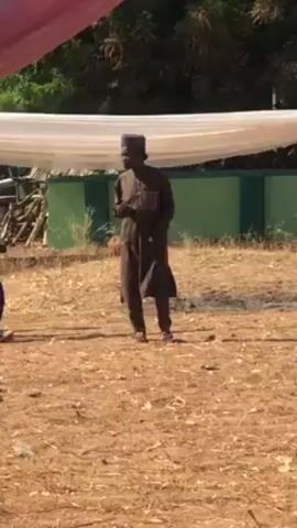The video shows a person wearing a brown robe and a matching hat walking towards the camera. The person is on a dry, grassy field. In the background is a green and white building and white fabric hanging overhead, likely a tent or awning. The lighting is bright, indicating a sunny day. Another person sits on the ground to the side. The main person is moving and gesturing as if speaking or directing.