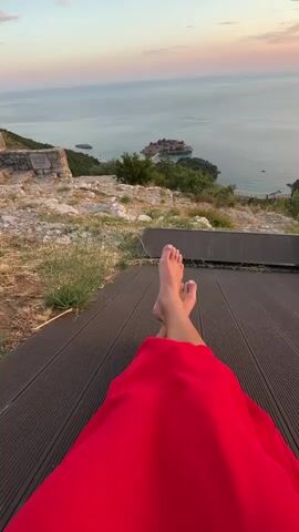 The video shows a first-person perspective of someone lying down on a wooden deck with their legs crossed. The person is wearing a long red skirt. Their feet are visible, and they appear to be barefoot. In the background, there is a scenic view of the ocean and a small island with buildings on it. The sky is a soft gradient of pink, orange, and blue, suggesting it is either sunrise or sunset. The foreground shows the textured wooden planks of the deck and some rocky terrain with sparse vegetation.
