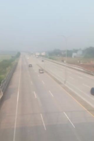 The video shows an aerial view of a highway with light traffic. The road is paved with multiple lanes and has white lane markings. There are a few cars and one truck visible on the highway. The surrounding environment appears to be rural, with some vegetation and a body of water visible in the distance. The sky is hazy, suggesting poor visibility. The video is shot from a high angle, looking down on the highway.