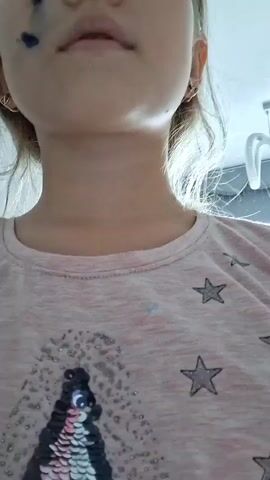 The video is a close-up shot of a young girl's face, focusing on her mouth and neck. She is wearing a light pink t-shirt with a sequined penguin design and scattered star motifs. The girl appears to have some dark marks on her cheek, possibly from makeup or paint. Her mouth is slightly open, and she seems to be speaking or singing. The lighting suggests it might be daytime, with natural light coming from the side, casting shadows. The background is blurred, but hints at an indoor setting, possibly a home environment. The overall mood is playful and candid. There is no visible text in the video.
