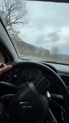 The video, shot from inside a car, depicts a first-person perspective of someone driving on a rural road, likely in a hilly or mountainous area. The dashboard, steering wheel, and interior of the car are visible. The weather appears cloudy and overcast. Trees and vegetation line the roadside. The road itself appears to be asphalt.