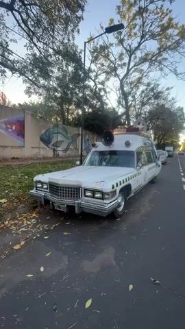 The video shows two parked cars on a street.  The first car is a white Cadillac, designed to resemble the Ghostbusters' Ecto-1 vehicle, and is parked near a low retaining wall with a mural on it.  The second car, parked directly behind the Cadillac, is a dark blue station wagon.  The street is lined with trees, and there are leaves on the ground.  The camera appears to be held by someone walking past the cars.