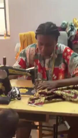A woman with cornrow braids is operating a treadle sewing machine at a yellow table. She is wearing a colorful, patterned shirt. In front of her is patterned fabric, and to the left are small tools. Behind her is a window and some other items. A small child is partially visible in the lower left corner of the frame. The scene appears to be indoors, possibly a workshop or tailoring space.