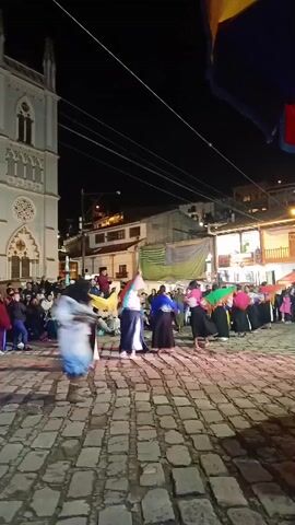 At night, a group of women in traditional clothing dances in a cobbled plaza, surrounded by spectators. The dancers wear long black skirts, white blouses, and colorful shawls. They move in a circle, spinning and waving their shawls. A large, gothic-style church stands to the left, illuminated by streetlights. Other buildings surround the plaza, some with lighted windows and balconies. A flag hangs in the foreground, partially obscuring the view. The atmosphere is lively and festive.