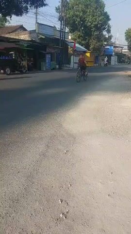 A boy rides a bicycle down a paved road. He is wearing a maroon t-shirt and black shorts. The boy is in the middle of the road and is pedaling. The sun is shining and there are buildings on the left side of the road. There are also a few motorcycles on the left side of the road.