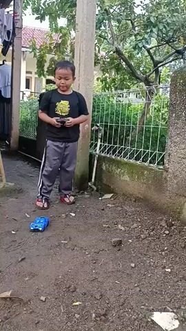 A young boy stands in an outdoor space, operating a remote-controlled blue toy car that rests on the ground in front of him. The boy is wearing a black t-shirt, grey sweatpants with white stripes, and sandals. He holds the remote control in his hands, focusing on the car. The ground is dirt-covered. A concrete pillar is situated behind the boy, with a fence, greenery, and a partially visible house in the background. Laundry hangs from a nearby structure on the left.  The video is shot from a stationary position.