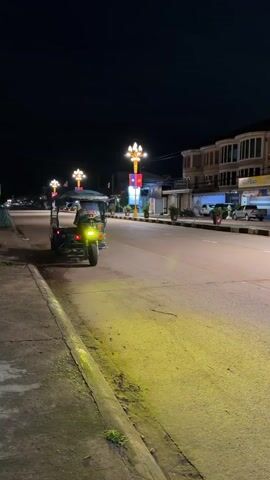 A tuk-tuk is parked on the side of a road at night. The tuk-tuk is blue and white and has a driver in it. The driver is wearing a dark jacket and smiling at the camera. The road is well-lit by streetlights and the headlights of the tuk-tuk. There are buildings on the other side of the road, and a few cars are parked there. The sky is dark, and the overall atmosphere is calm and peaceful.