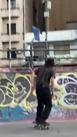 A young male skater performs a trick on a skateboard in a concrete skatepark area with graffiti art on its walls. He wears dark clothing and a cap. The skateboard is gray and has visible wheels. The skateboarder accelerates up a curved section of the concrete wall, appears to slightly jump, and then returns to riding the skateboard. The background reveals an urban environment with buildings and street signs. The light is somewhat diffused, indicating an overcast day.
