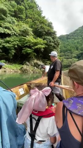 A small group of people are on a boat navigating a narrow river surrounded by lush green forested hills. The boat appears to be a traditional wooden vessel. Two men are actively engaged in propelling the boat. One man, wearing a black t-shirt, shorts, a white baseball cap, and sunglasses, stands at the stern, using a long pole to push off the riverbed. He is smiling and looking towards the front of the boat. The other man, wearing a white t-shirt and a dark baseball cap, is leaning over the side of the boat near the front, also seemingly involved in maneuvering the boat. The passengers are seated behind the two men. In the foreground, partially visible, are the backs of several passengers. One passenger in the center is wearing a red life vest and has a large pink bow tied to the back of their head. Another passenger to the right is wearing a patterned garment and a hat. The water is a calm, green color. The sky is partly cloudy. The overall impression is a scenic boat trip through a natural environment.