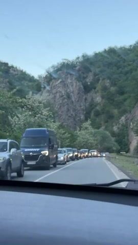 The view from inside a car shows a line of vehicles stopped on a road. On the side of the road is a lush green hillside, capped with a prominent rocky cliff. The sky is mostly clear, pale blue. The vehicles in the queue range from vans to cars, and a pickup truck is also visible. All vehicles are facing the same direction, implying a traffic jam.