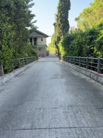 A paved road ascends between lush green trees and foliage. On the left, a light-colored two-story building with a green roof and balcony is visible amidst the trees. The road is bordered by concrete walls with metal railings on both sides. In the distance, a white barrier is placed across the road, partially blocking it. The sky is clear and bright, suggesting daylight.