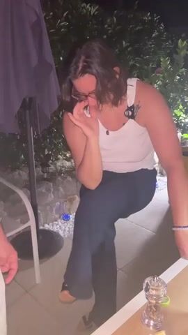 A person with short brown hair, wearing glasses, a white tank top, and dark pants, is bending over with their face in their hands. They are outdoors at night, with a table, chairs, and plants visible in the background. There is a salt and pepper grinder on the table.