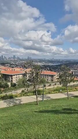 The video shows a panning shot of a city skyline from a grassy hilltop on a cloudy day. In the foreground, there is a manicured lawn and several young trees. In the midground, there is a paved walkway with streetlights and more trees. Beyond the walkway, there are apartment buildings with red tile roofs. In the background, the city skyline is visible with taller buildings in the distance, and a body of water, likely a strait or bay. A minaret of a mosque is visible on the right side of the frame. The shot is taken from a high vantage point, offering a panoramic view of the urban landscape.