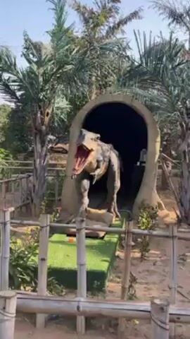The video shows an animatronic Tyrannosaurus Rex dinosaur. The dinosaur is standing on a rock-like base in front of a concrete-looking tunnel. The T-Rex is roaring with its mouth wide open, showing its teeth. The background includes green foliage and what looks like a wooden fence. The scene appears to be part of a theme park or a similar attraction.