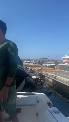 The video shows several men on a boat during a sunny day. The scene opens with a man in a green long-sleeve shirt, sunglasses, and patterned shorts, wearing a fanny pack, standing on the deck. The clear blue sky and other boats suggest a marina or open water setting.

The main activity involves two men fueling the boat. One man, dressed in a white short-sleeve shirt and patterned shorts, is carefully pouring liquid from a dark green jerrycan into a fuel intake on the boat's floor. Another man, wearing a red t-shirt and striped shorts, stands beside him, observing the process. A fourth man, seated in the foreground, is visible wearing a black t-shirt with "LONSDALE LONDON" printed on it and pink shorts. He is holding a smartphone and watching the refueling.

The weather is bright and clear, with a vivid blue sky. The side of the boat features the text "CRETAN Meraya" and a blue cleaning brush is hanging. The overall atmosphere appears calm and focused on the task at hand.
